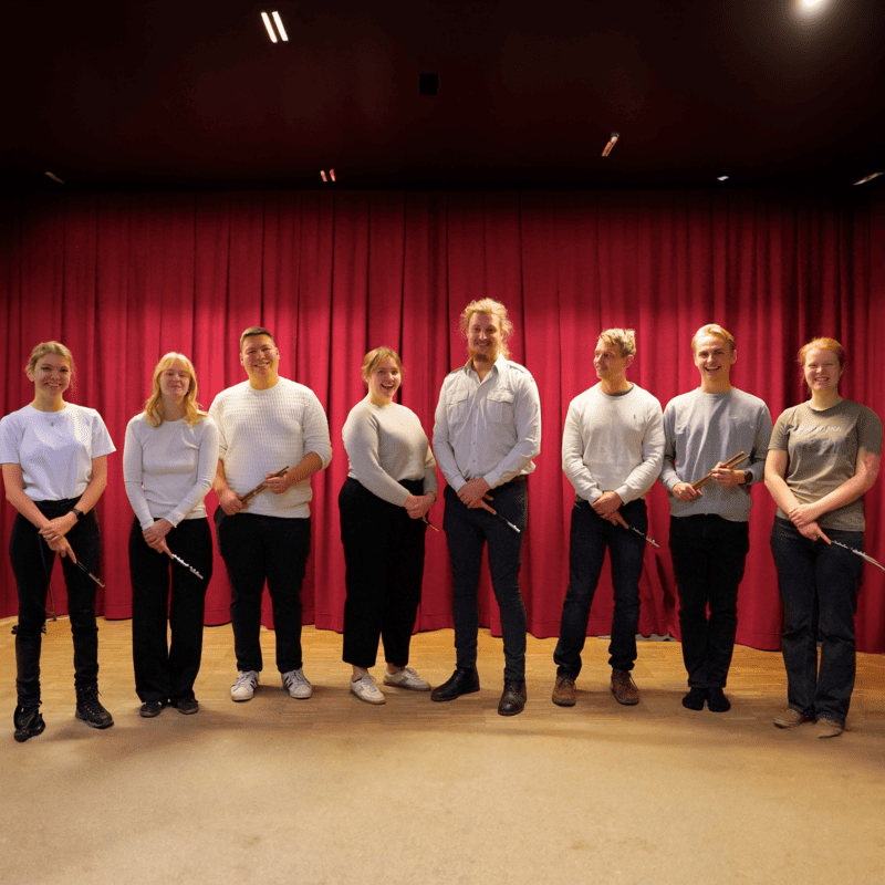 Otte smilende medlemmer fra Det Ny Fynske Tambourkorps står på en scene foran et mørkerødt gardin.